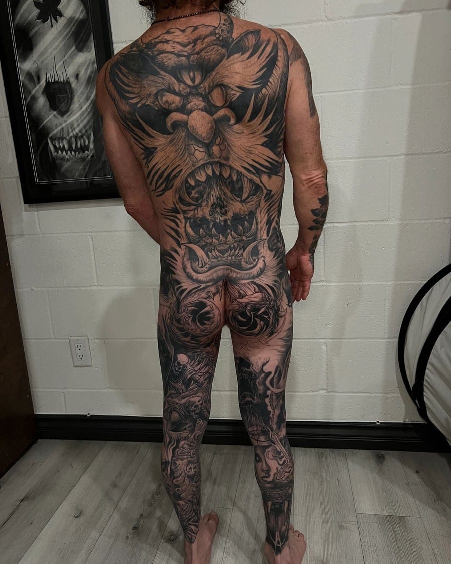 Dense black and grey illustrative tattoo of monster faces and skulls across a back and legs