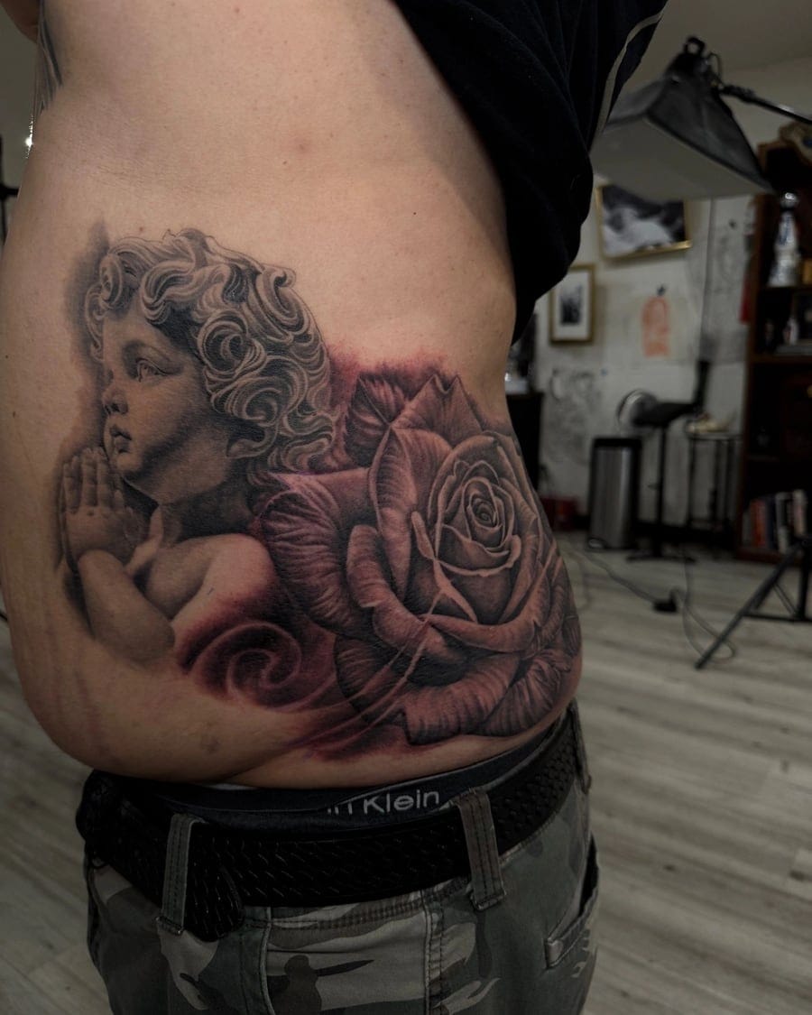 Black and grey tattoo of a cherub in prayer beside a large shaded rose on the stomach