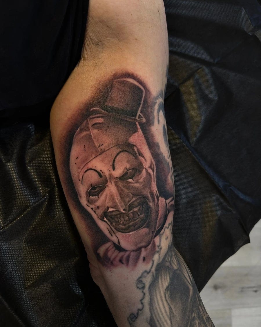 Black and grey forearm tattoo of a clown face with a hat and ruffled collar