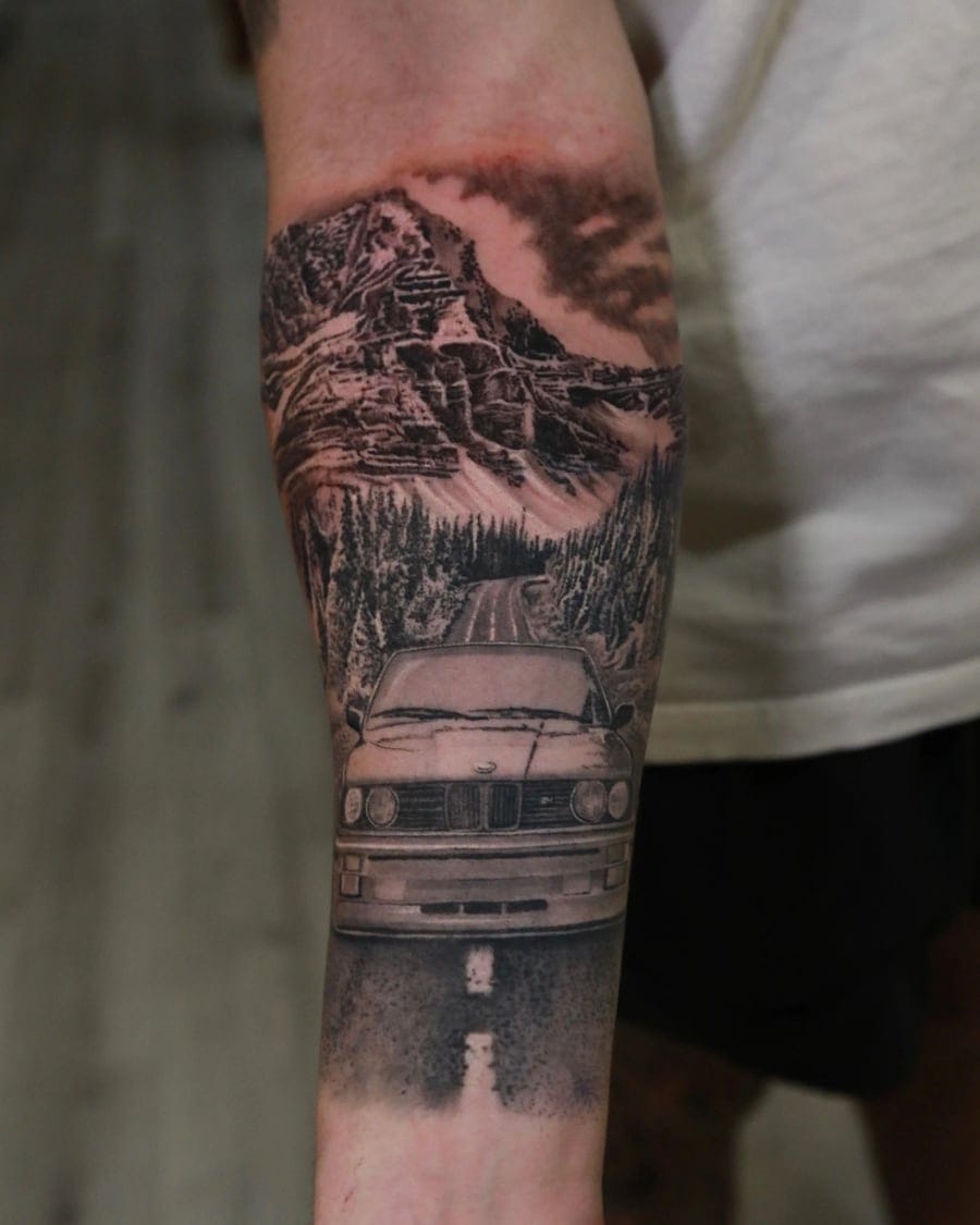 Black and grey forearm tattoo of a car on a forest road below snow-capped mountains