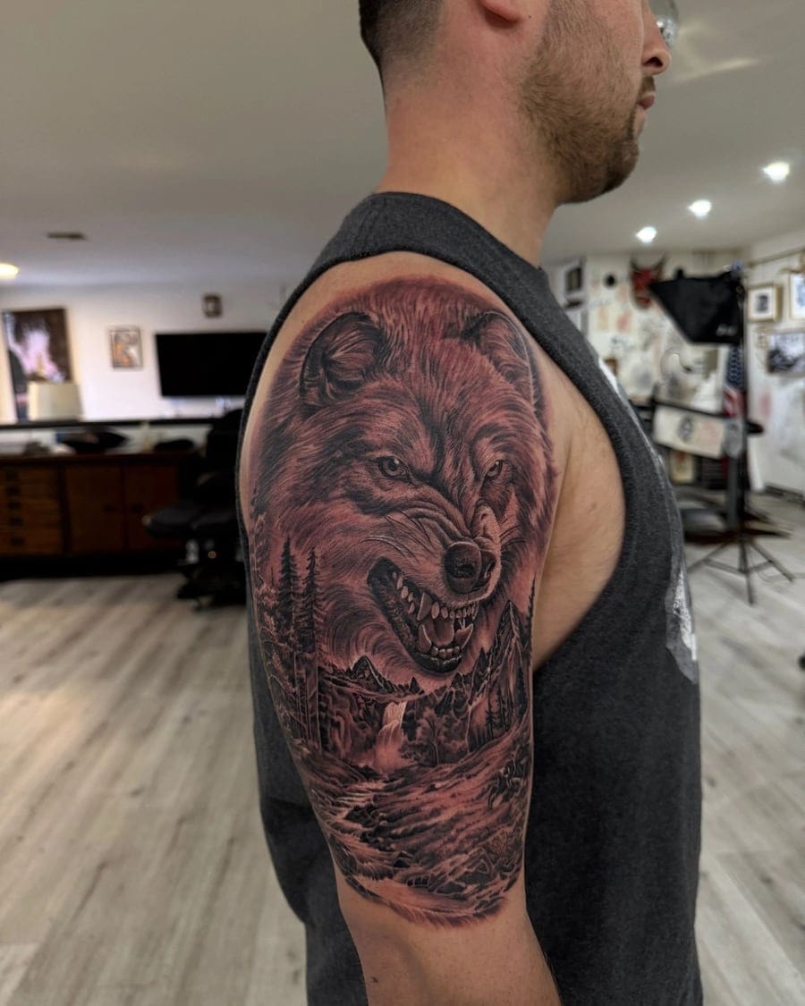 Black and grey upper arm tattoo of a snarling wolf above a forest waterfall scene