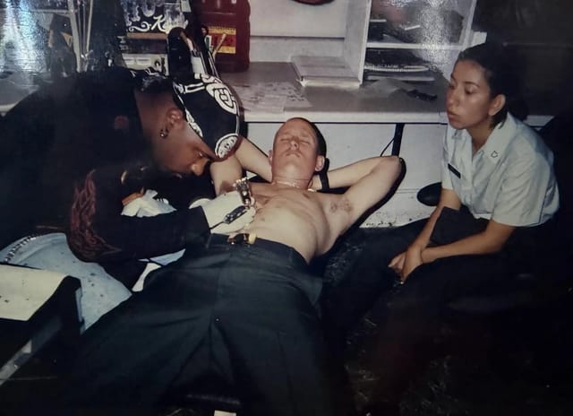 A young Cris Skiver, then an enlisted soldier, lying back while a fellow soldier tattoos his chest.